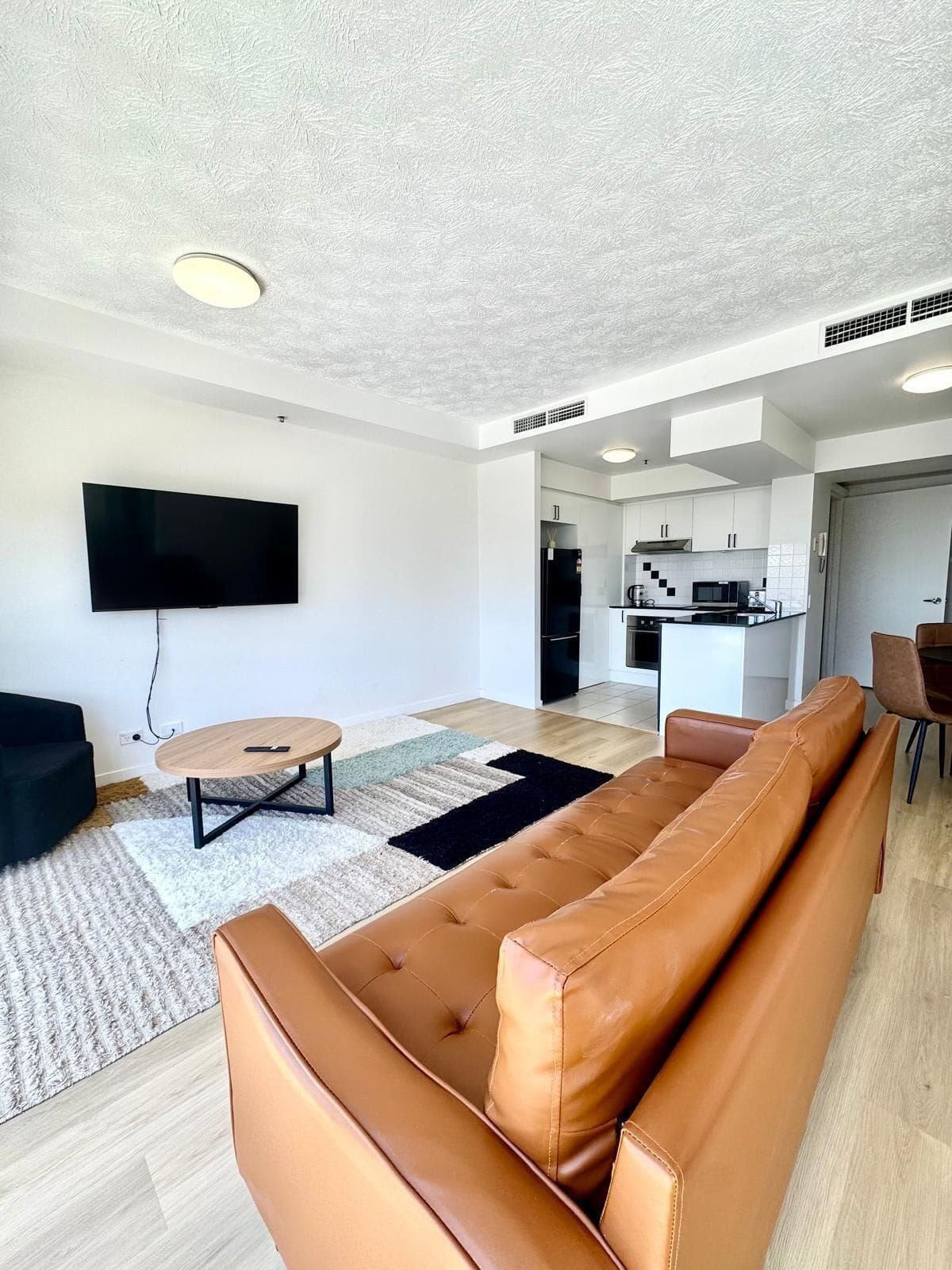 A modern living room with a large brown leather sofa, a flat-screen TV mounted on the wall, and a small round coffee table.