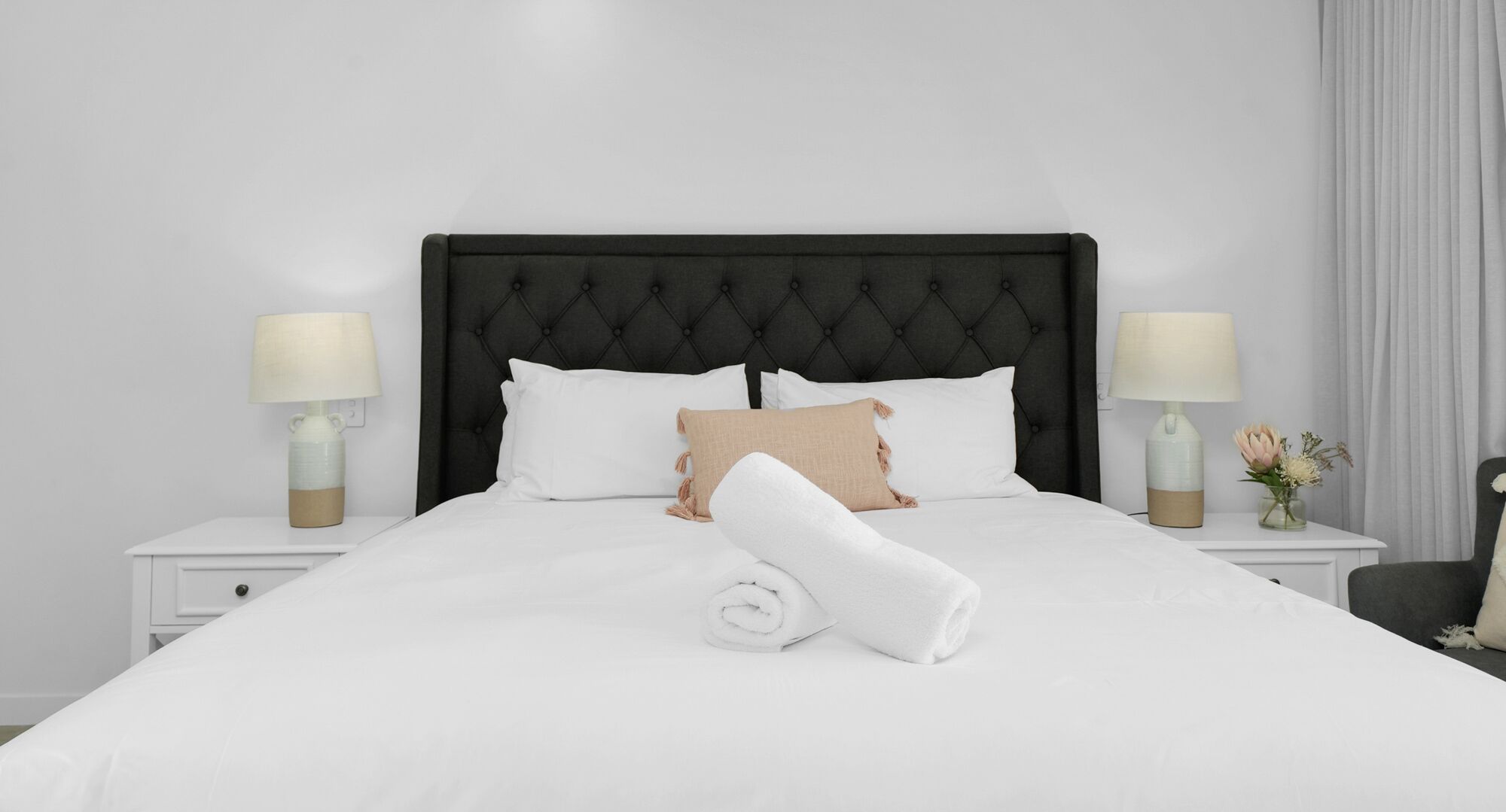 Un lit bien fait, avec du linge de lit blanc, une tête de lit noire à motifs et deux tables de chevet ornées de lampes et d’un vase de fleurs.