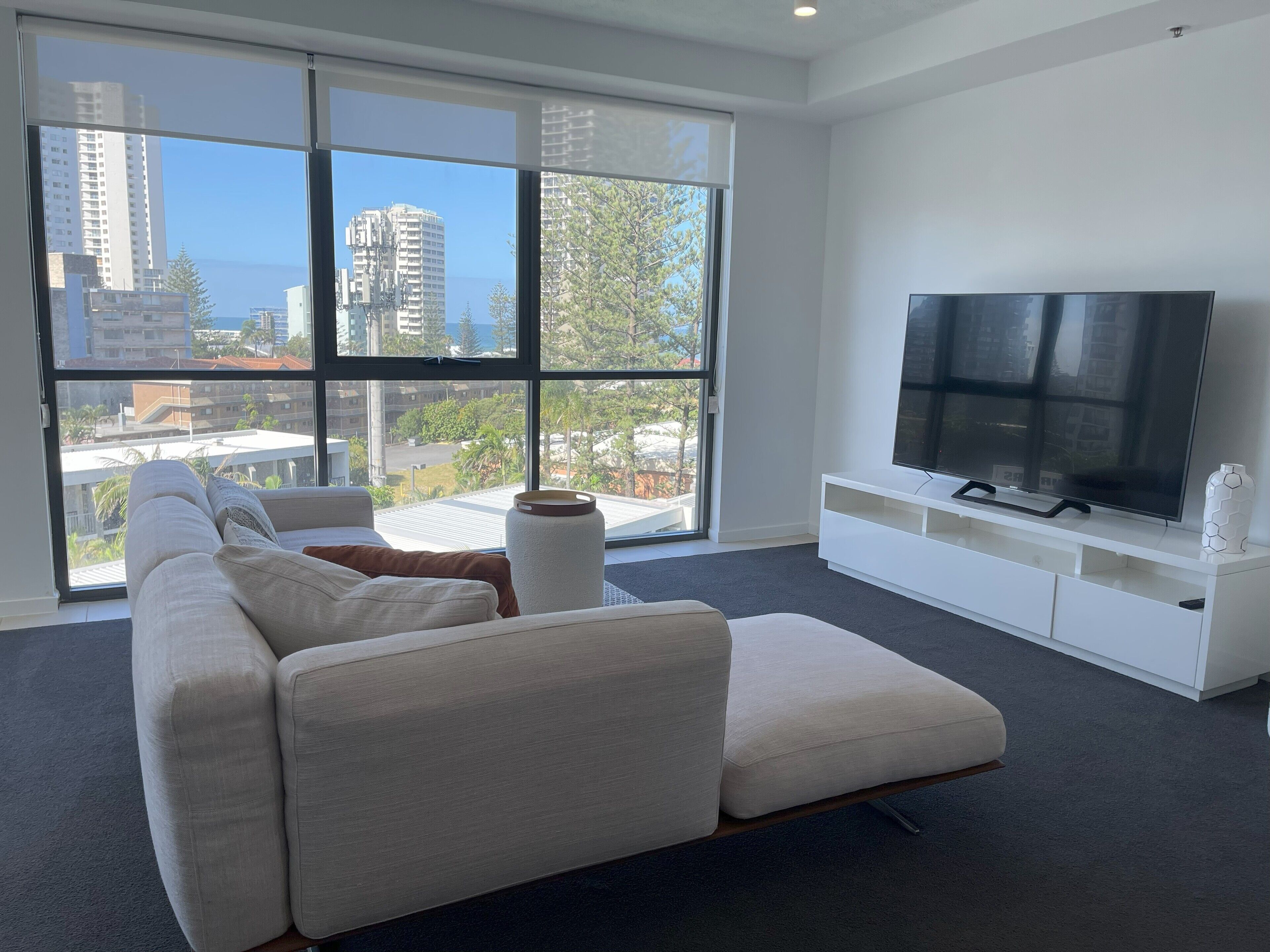 A modern living room with a large window, a white sofa, a flat-screen TV, and a minimalist white entertainment unit.