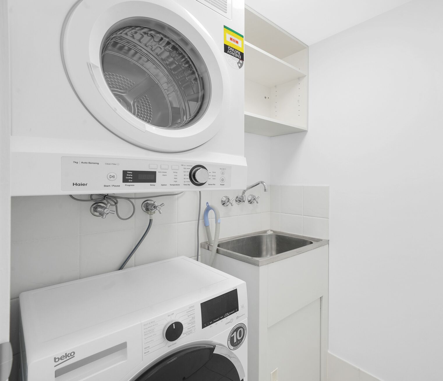 A compact laundry area with a front-loading washer and dryer, a small sink, and a shelf above the washer.