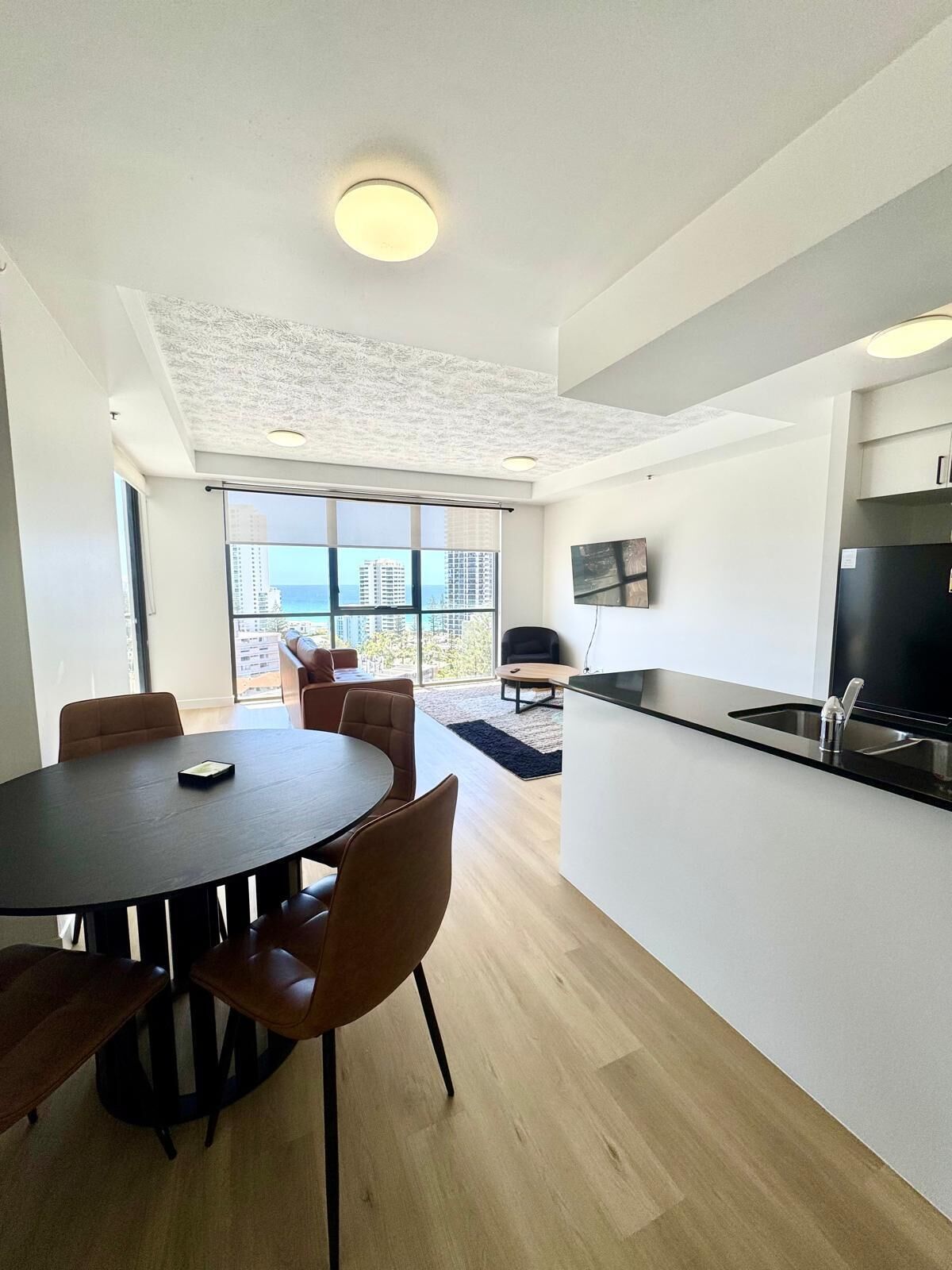 A modern kitchen with a black countertop and stainless steel appliances, a dining area with a round table and chairs, and large windows offering a view of the city and ocean.
