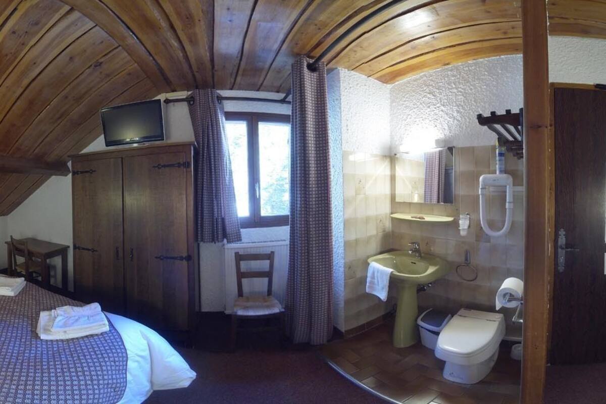 Een slaapkamer met een bed, houten plafond, televisie, een klein tafeltje en een badkamer met wastafel en toilet.