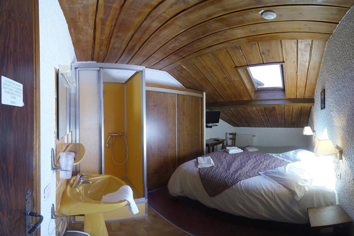 Een kamer met een houten plafond, een bed, een klein bureau, een televisie en een badkamer met een geel wastafelmeubel en douche.
