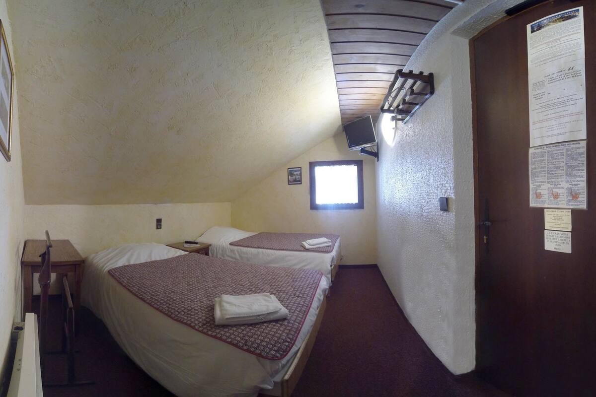 Een kleine, zwak verlichte kamer met twee bedden, een houten bureau en een raam.