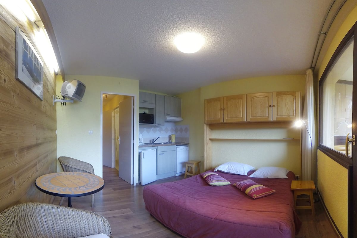 Een kleine kamer met een bed, een kitchenette, een eettafel en een stoel.