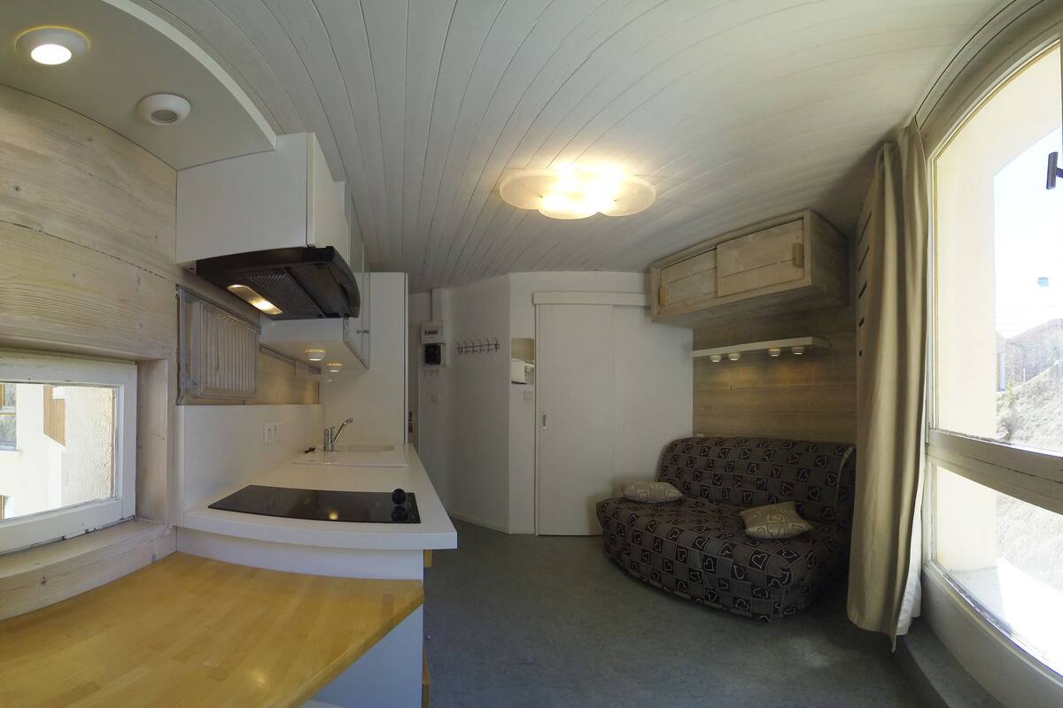 Een compacte kamer met een gebogen houten plafond, een inbouwspoelbak, een bank en een raam met gordijnen.