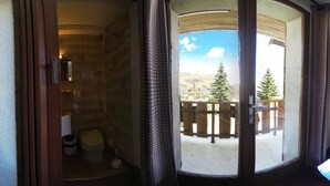 Habitación de hotel con cama, escritorio, silla, televisor y balcón con vistas a árboles y edificios.
