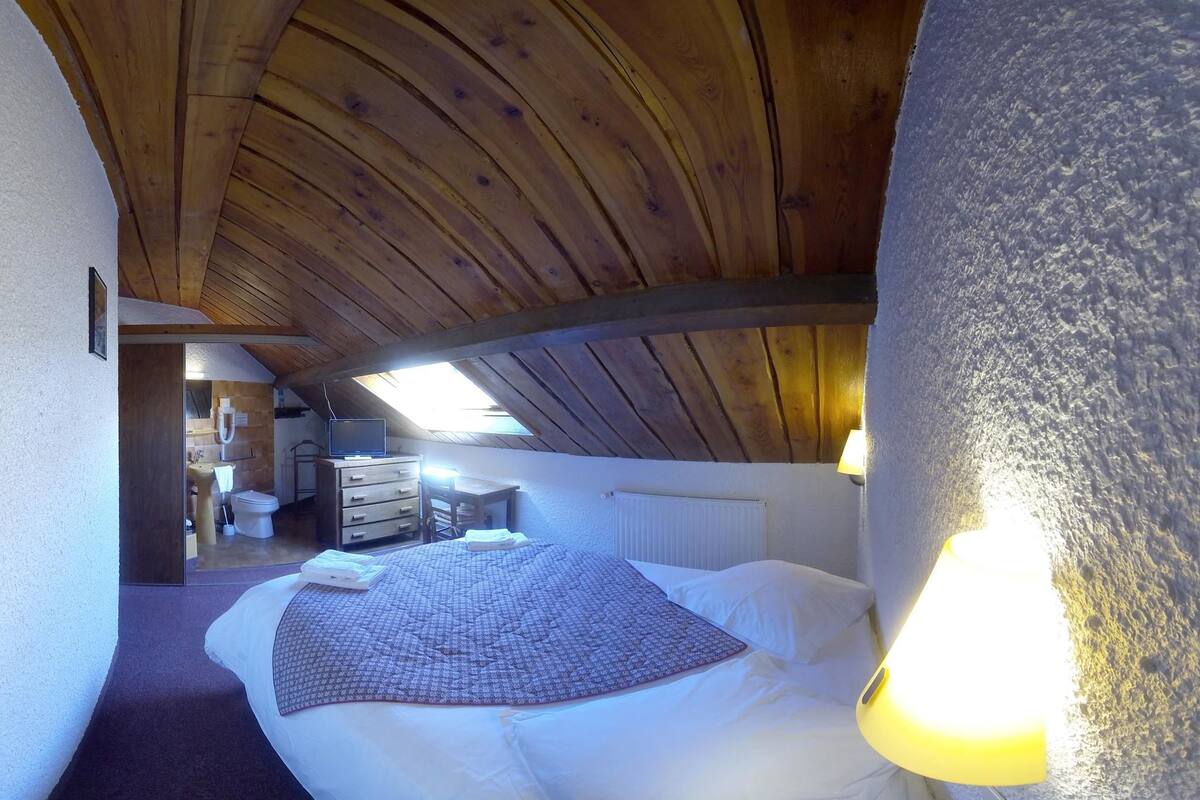 Een slaapkamer met een houten plafond, een bed met witte lakens, een bedlampje en uitzicht op een badkamer en een keukenhoek.