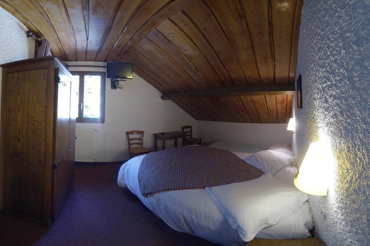 Een slaapkamer met een schuine kap, een bed met een geborduurde deken, twee stoelen, een klein tafeltje en een aan de muur bevestigde televisie.