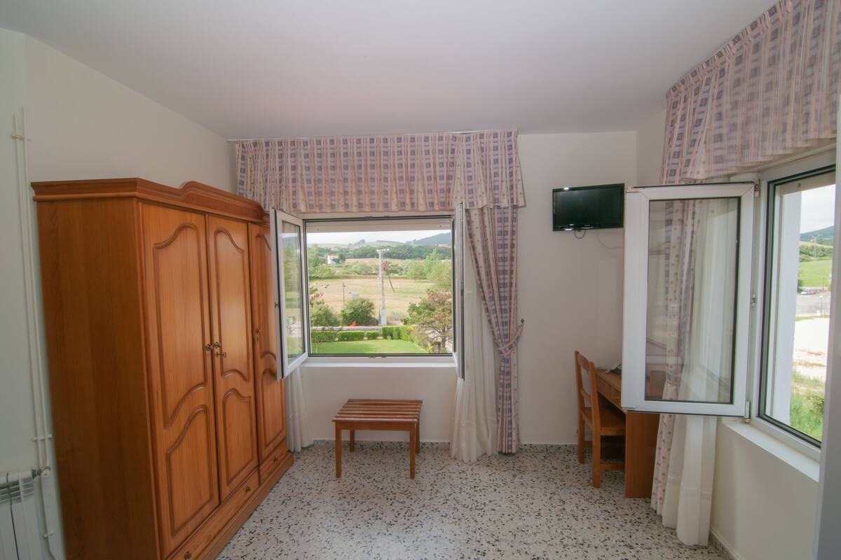 A room with a large window, a wooden wardrobe, a small wooden bench, and a television mounted on the wall.