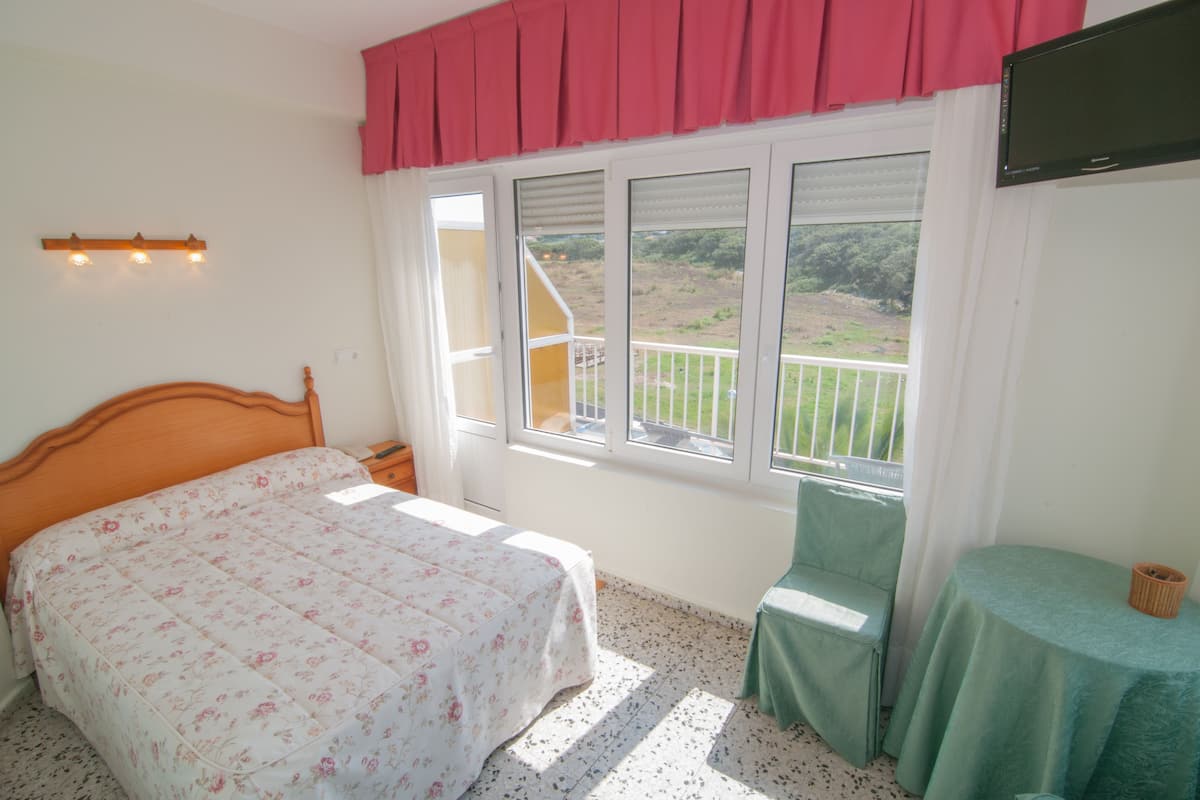A bedroom with a bed, a chair, a table, a TV, and a window with a view of a balcony and greenery.