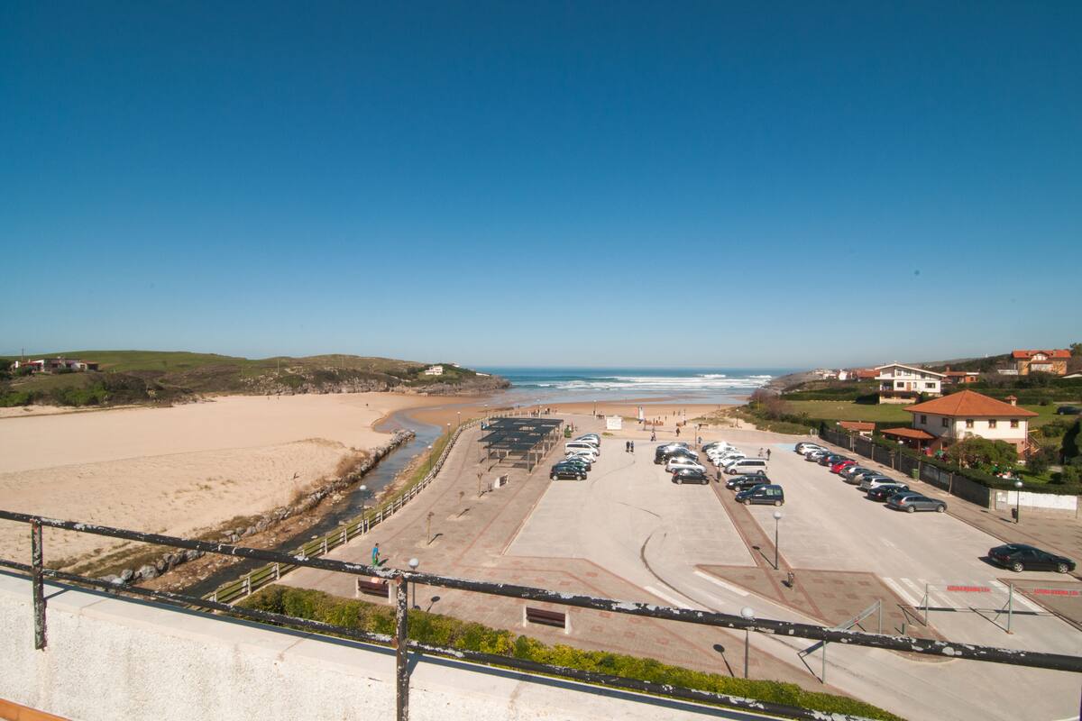 A coastal road with parked cars, a beach, and houses in the background.