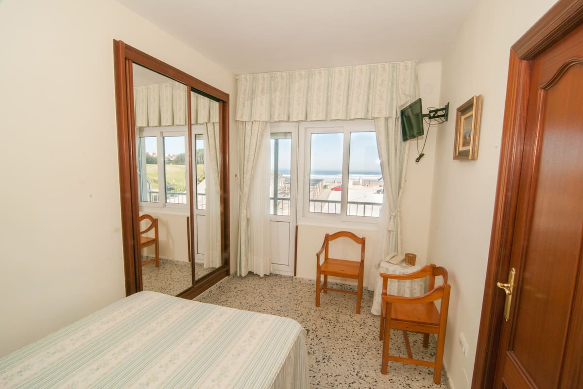 A bedroom with a bed, a chair, a small table, a window with curtains, a door, and a wall-mounted TV.