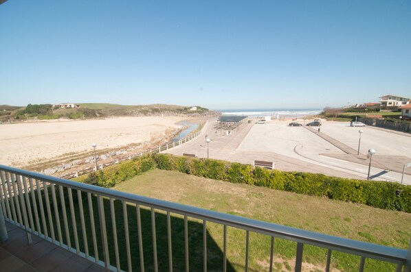 Vista costera desde un balcón con vista a una playa, un estacionamiento y una carretera, bajo un cielo despejado.
