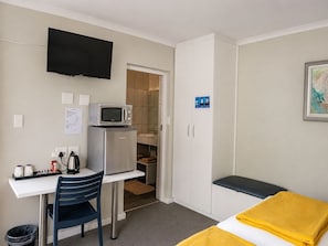 A compact hotel room with a kitchenette, a television mounted on the wall, a bed with a yellow blanket, and a single chair.