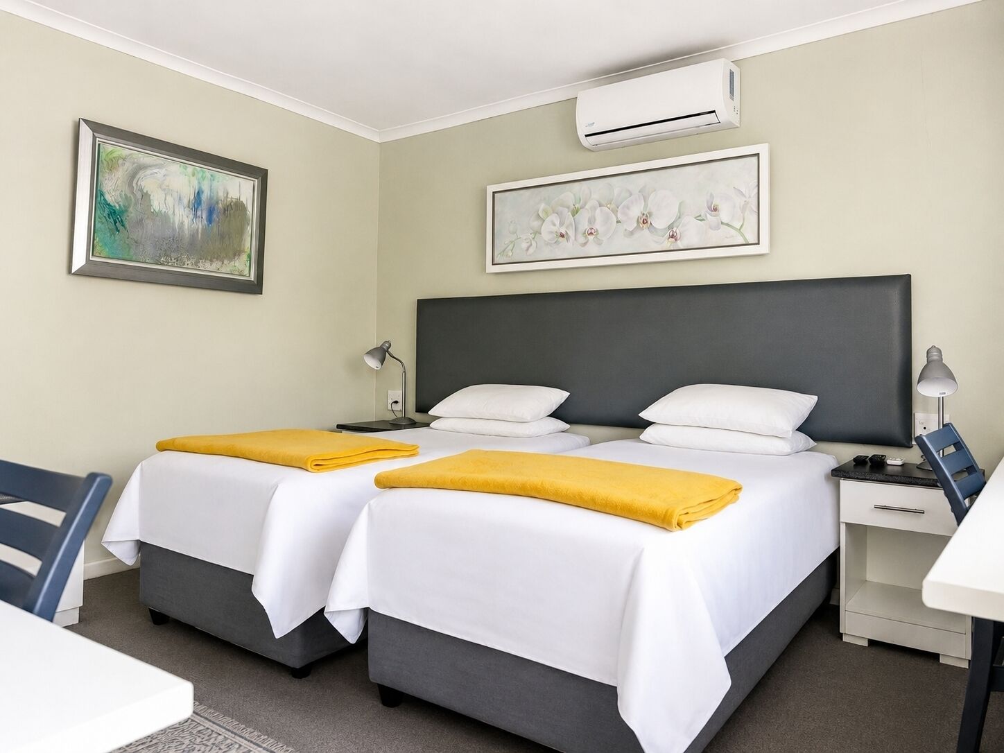 Quarto de hotel com duas camas, uma mesa, uma cadeira, ar-condicionado e obras de arte nas paredes.