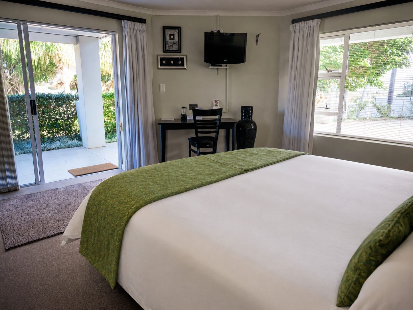 Habitación de hotel con cama, escritorio, silla, televisor y vista a zonas verdes.