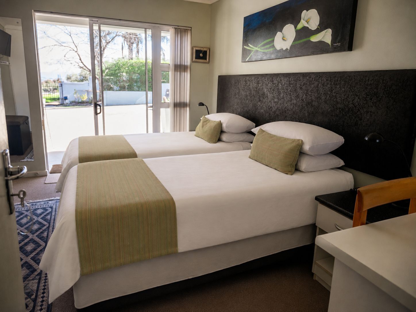 A hotel room with two beds, a desk, a chair, a painting, and a view of the outdoors.