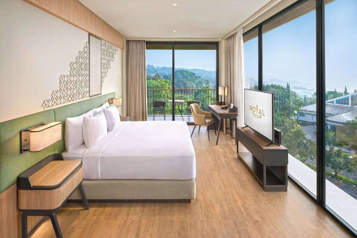 Ein modernes Hotelzimmer mit einem großen Bett, einem Schreibtisch, einem Fernseher und einem Balkon mit Blick auf die Stadt.