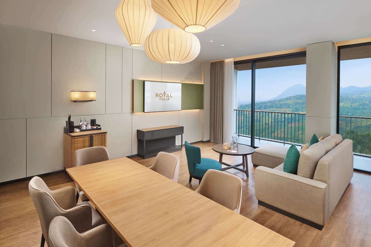 Ein Hotelzimmer mit einem Holzespeisetisch, beigen Sofas, einem kleinen runden Tisch mit Stühlen, einem an der Wand montierten Fernseher und einem Balkon mit Bergblick.