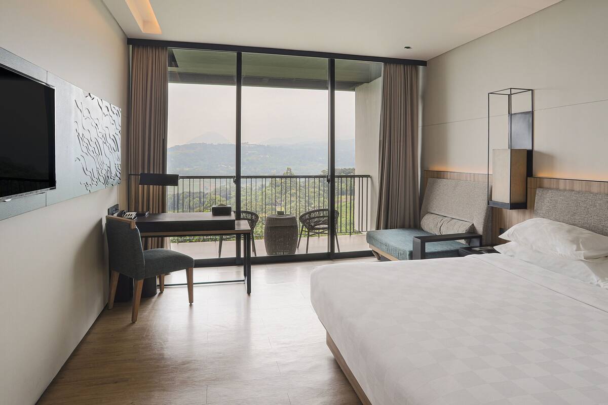 Ein Hotelzimmer mit einem großen Bett, einem Schreibtisch mit Stuhl, einem kleinen Tisch und einer Couch. Ein Fernseher ist an der Wand montiert und es gibt einen Balkon mit Bergblick.