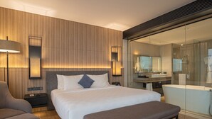 Une chambre d’hôtel moderne dotée d’un grand lit, de tables de chevet, d’un banc, d’un canapé, d’une salle de bain avec une douche à parois de verre et d’un grand miroir.