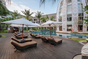 A poolside area with lounge chairs and umbrellas, surrounded by a wooden deck and a building with patterned exterior walls. - Golden Tulip Jineng Resort Bali (Kuta)