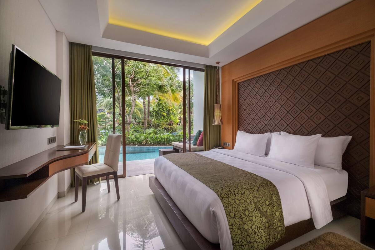 Ein Hotelzimmer mit einem großen Bett, einem Schreibtisch mit Stuhl, einem Fernseher und Blick auf einen Swimmingpool und Grünflächen.