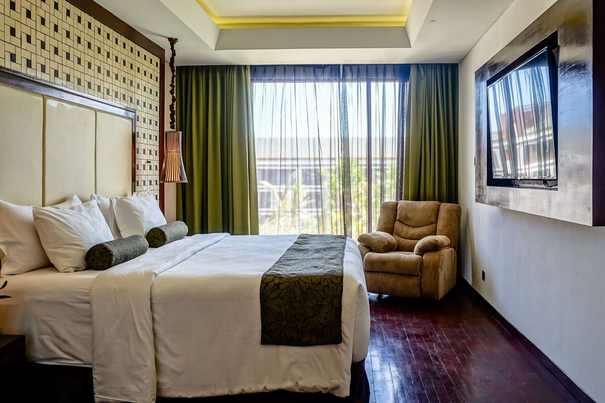 Une chambre d’hôtel avec un grand lit, une chaise, un téléviseur fixé au mur et une fenêtre avec des rideaux verts.