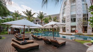 A poolside area with lounge chairs and umbrellas, surrounded by palm trees and a building with patterned windows. - Golden Tulip Jineng Resort Bali (Kuta)