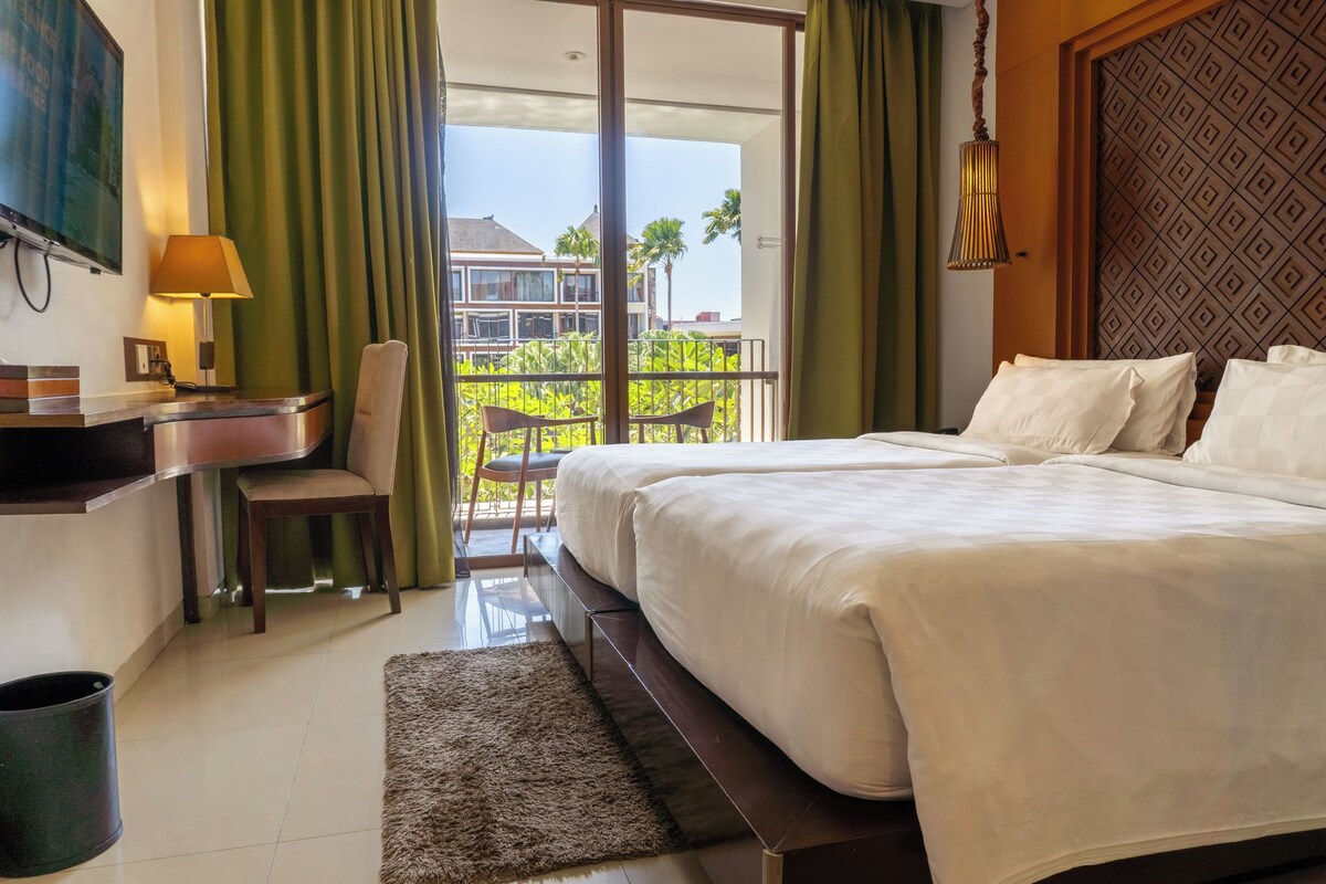 A hotel room with a bed, a desk, a chair, a TV, and a balcony with a view of buildings and palm trees.