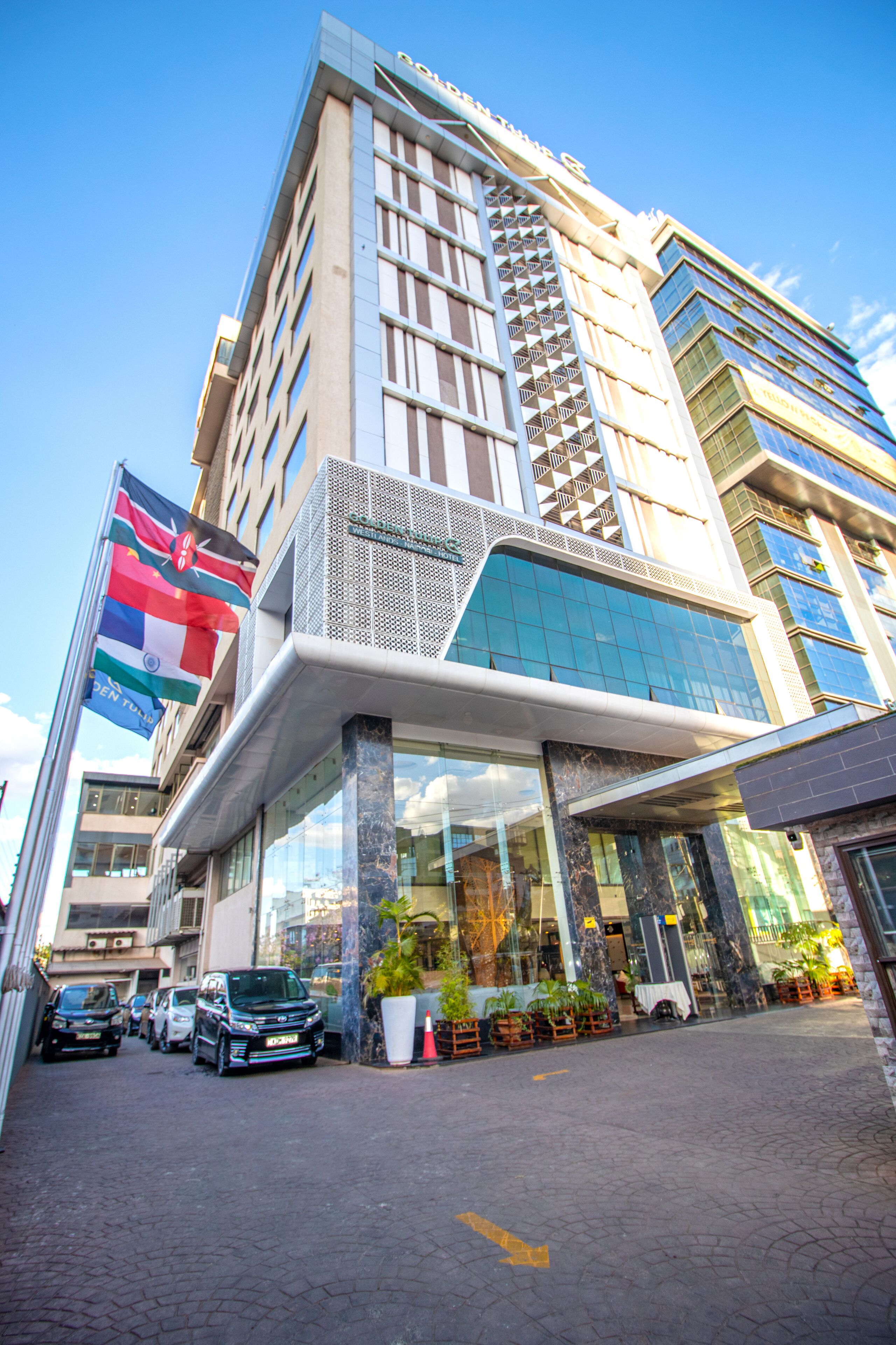 A modern hotel with a prominent glass facade, a covered entrance, and a flag on the left.