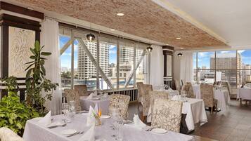 Restaurante com mesas postas para refeições, grandes janelas com vista para a cidade e teto com padrão texturizado.