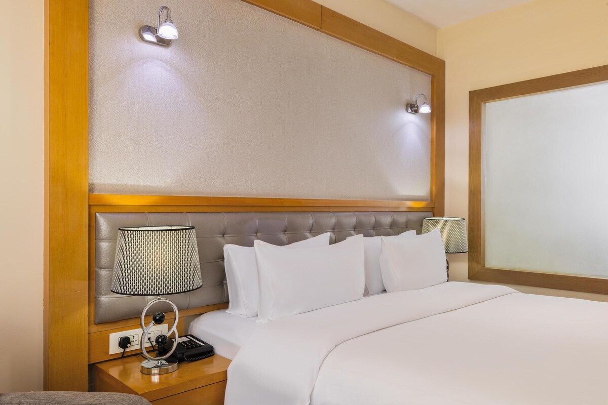 Ein Hotelzimmer mit einem großen Bett, zwei Nachttischlampen, einem Nachttisch mit Telefon und Lampe sowie einem Kopfteil mit Lichtleiste.