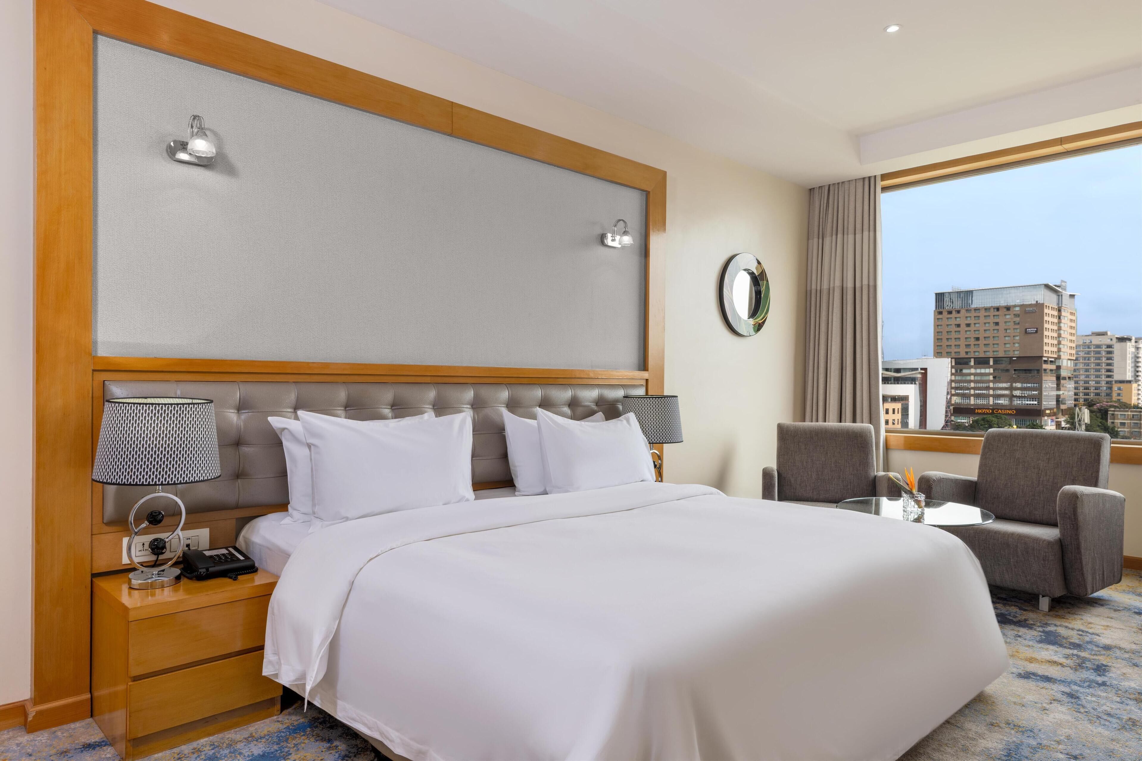 A hotel room with a large bed, a nightstand with a lamp, a chair, and a view of the city.