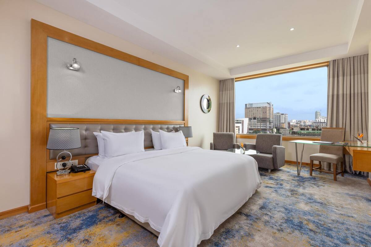 A hotel room with a large bed, a desk, and a chair, a view of the city, and a clock on the wall.
