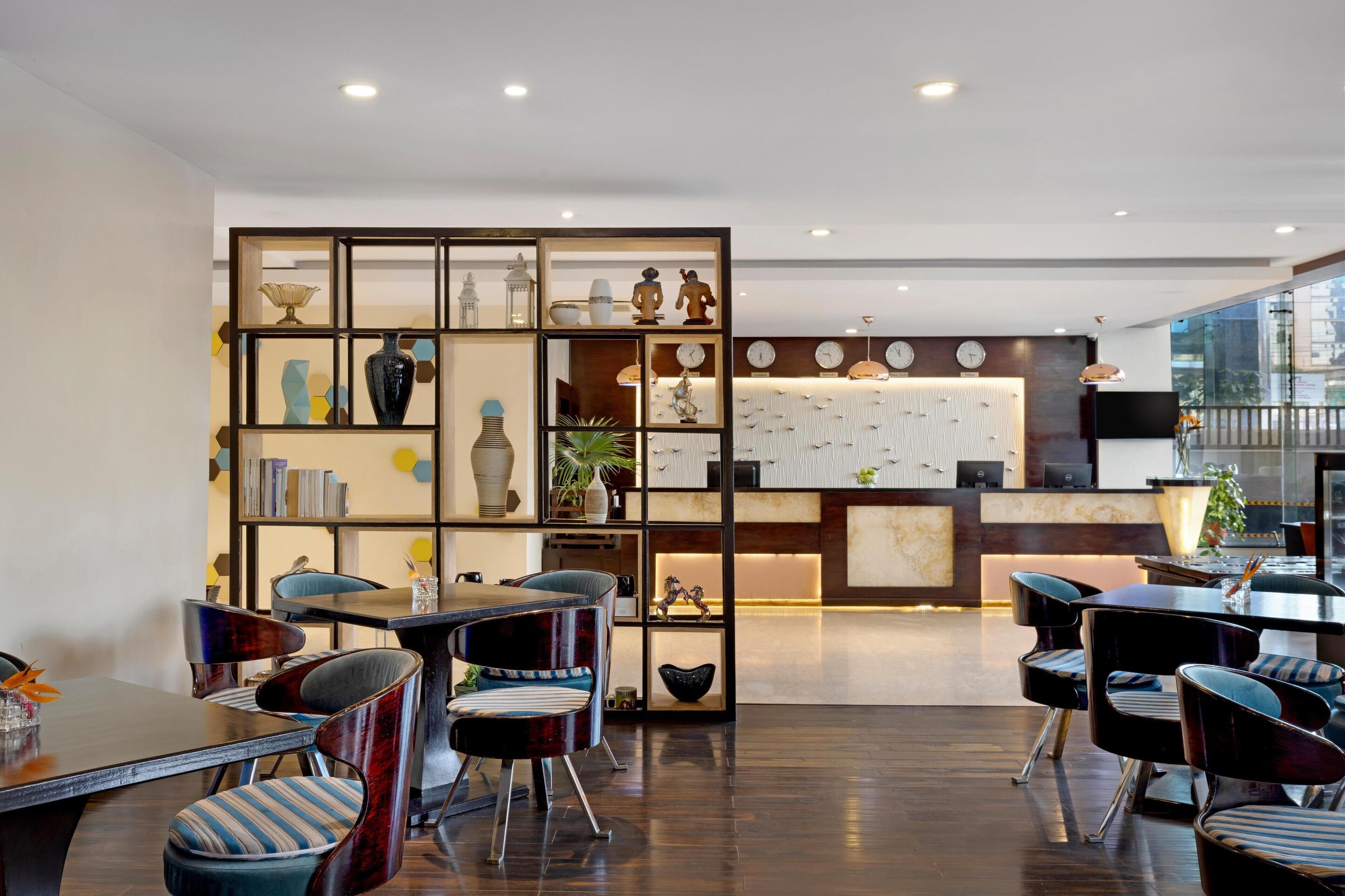 A modern hotel lobby with a reception desk, seating area with striped chairs, and a decorative wall with various items.