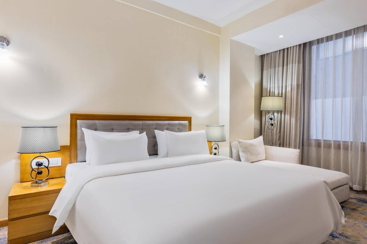 A hotel room with a large bed, two bedside tables with lamps, a headboard, and a window with curtains.
