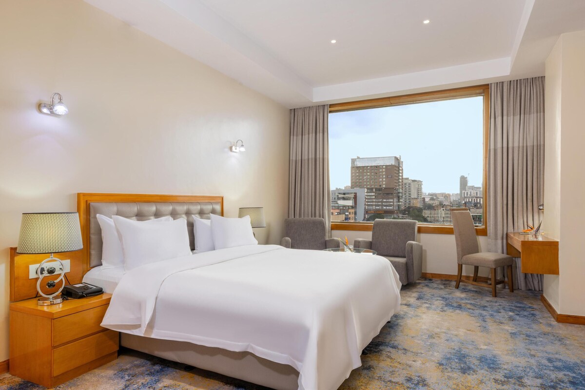 Ein Hotelzimmer mit einem großen Bett, einem Schreibtisch, zwei Stühlen, einem Nachttisch mit Lampe und Blick auf die Stadt durch ein großes Fenster.