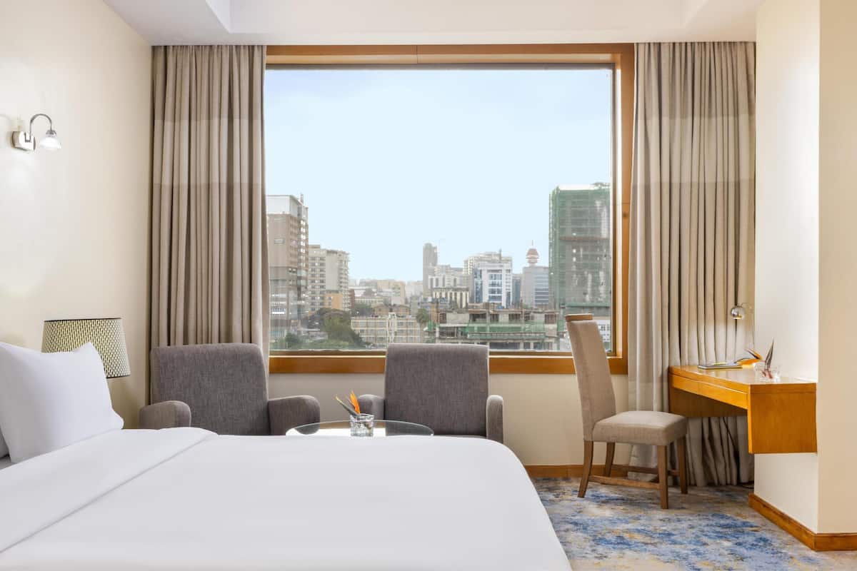 A hotel room with a large bed, two armchairs, a desk, and a view of a cityscape through a large window.