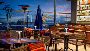 Eine Dachterrasse mit Korbmöbeln, einer Bar mit Flaschen und Blick auf die Stadt im Abendlicht.