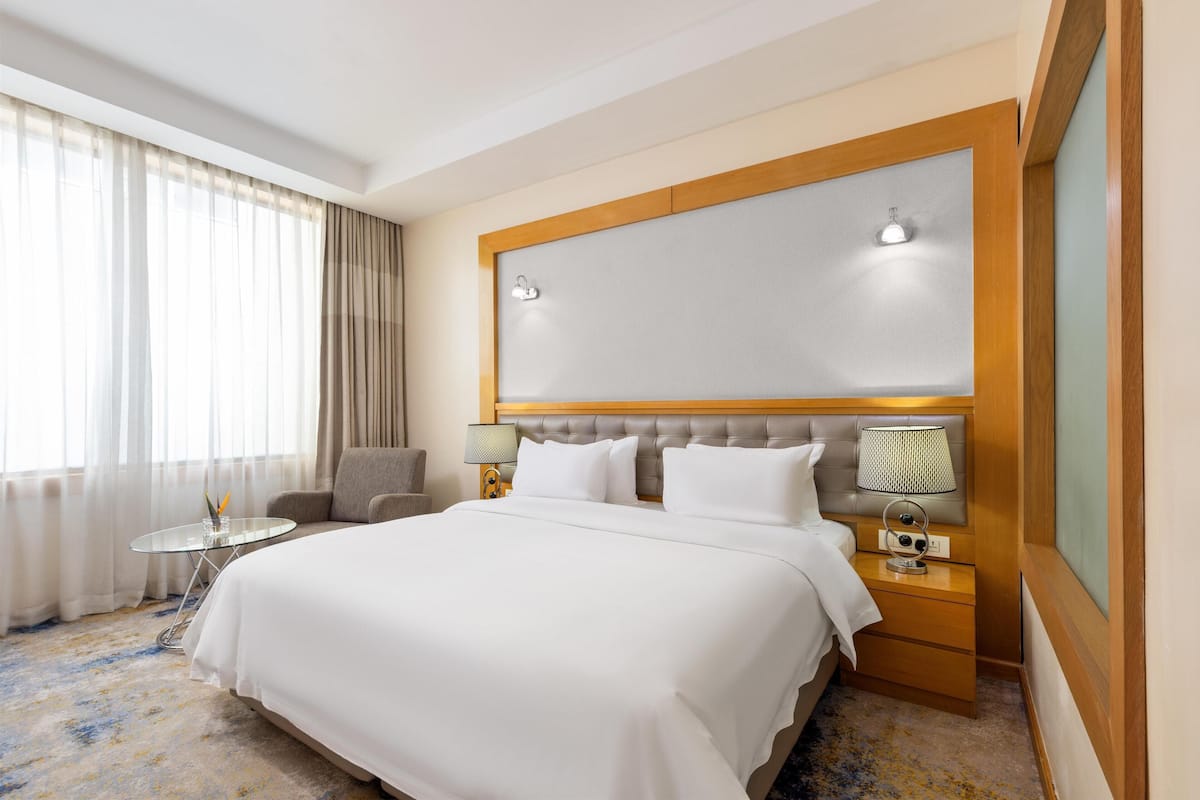 A hotel room with a large bed, a bedside table with a lamp, a chair, and a view of the outdoors through sheer curtains.