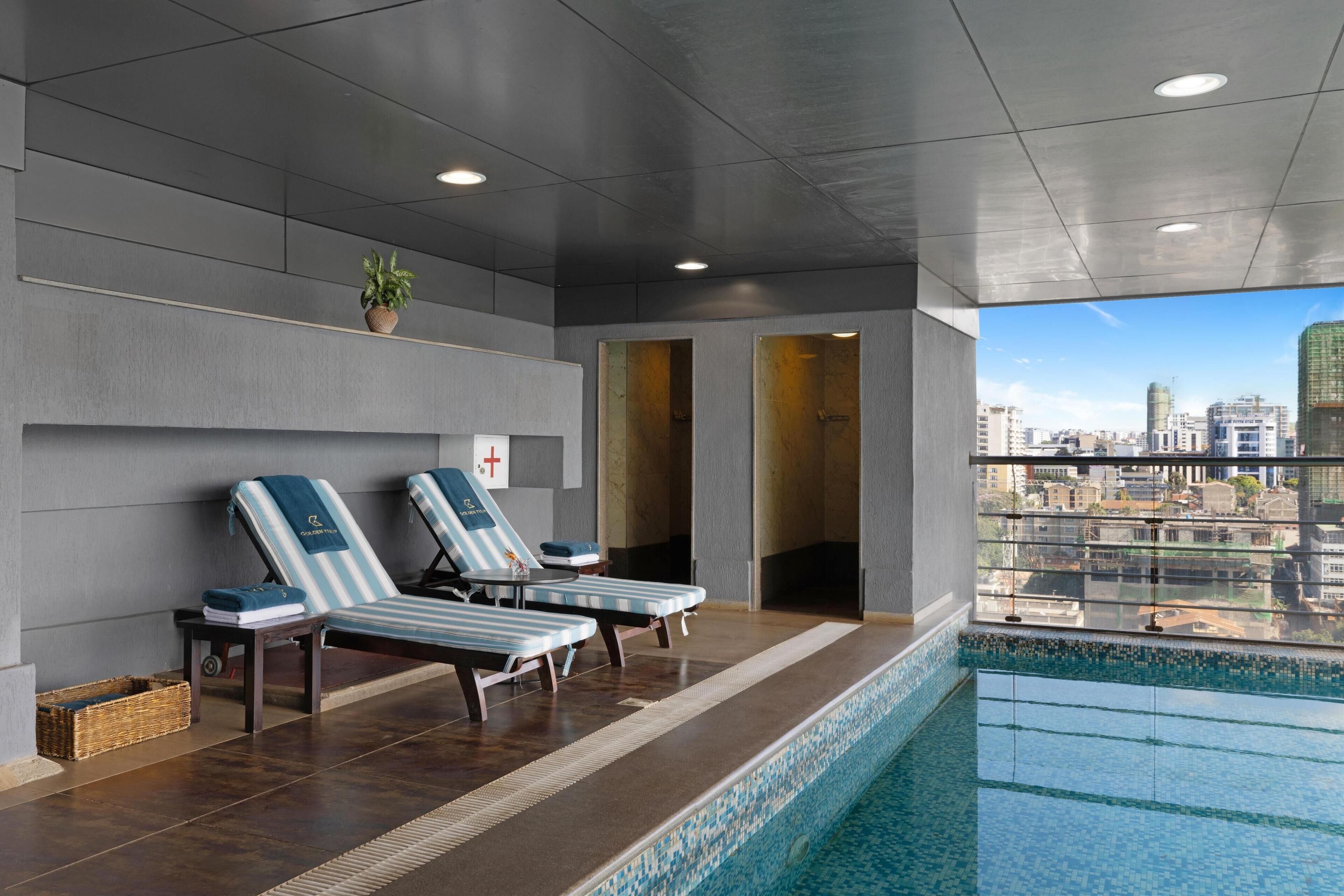 A modern indoor pool area with lounge chairs, a city view, and a tiled pool.