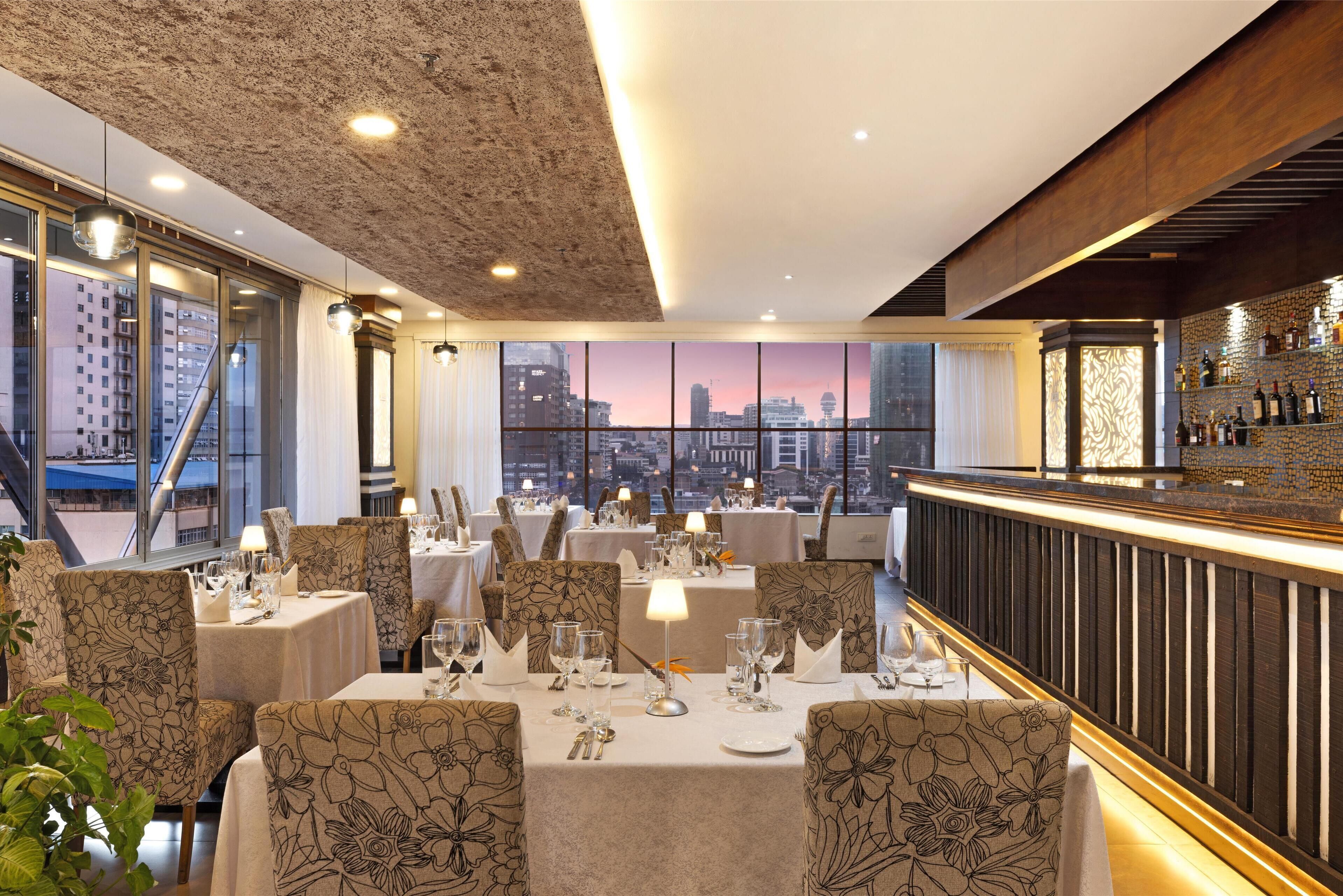 Un restaurante con mesas dispuestas para comer, una zona de bar y grandes ventanales con vistas a la ciudad.