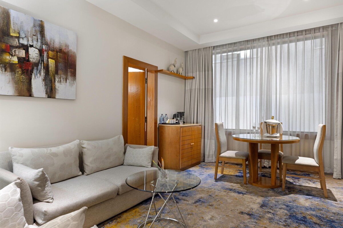 Ein modernes Wohnzimmer mit einer Couch, einem Glastisch, einem Holzesstzimmer mit Stühlen, einem Schrank und einem großen abstrakten Gemälde an der Wand.
