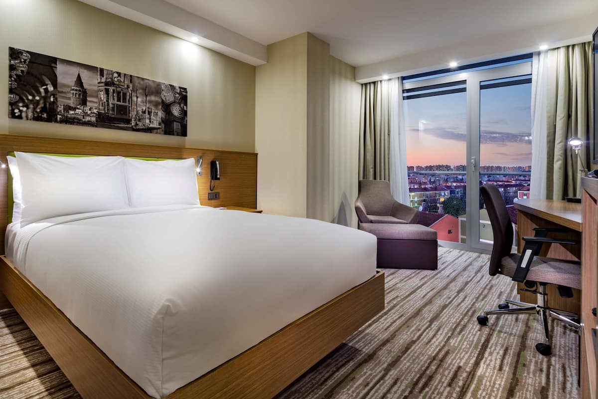 A hotel room with a large bed, a desk with a chair, a television, and a city view through the window.