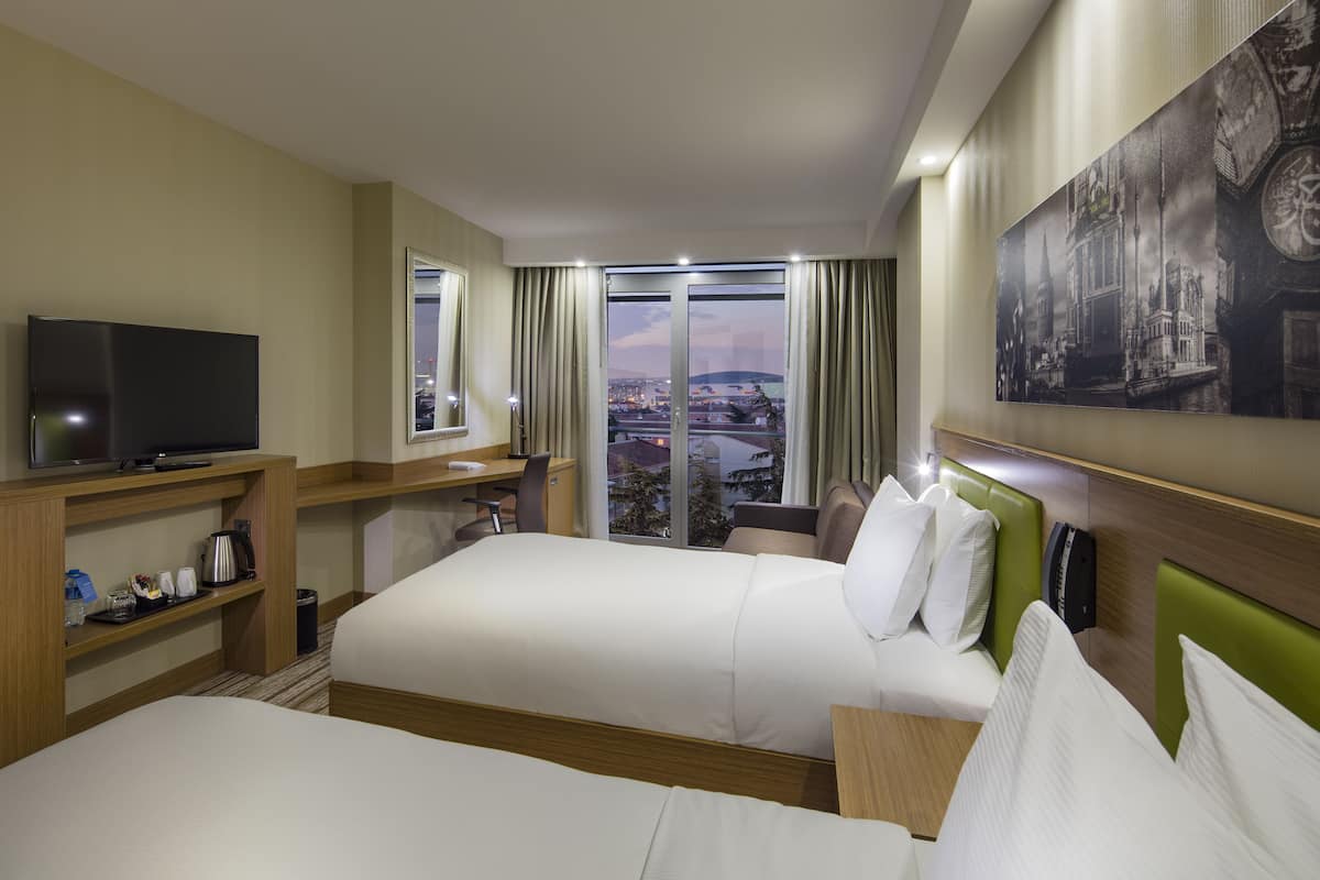 A hotel room with two beds, a television, a desk with a chair, and a large window with a city view.