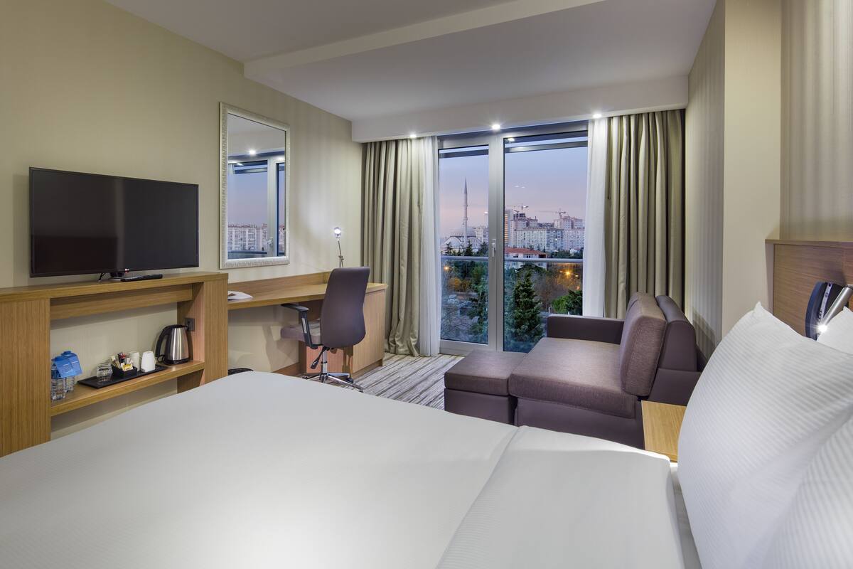 A hotel room with a bed, a desk, a chair, a sofa, a TV, and a large window with a city view.