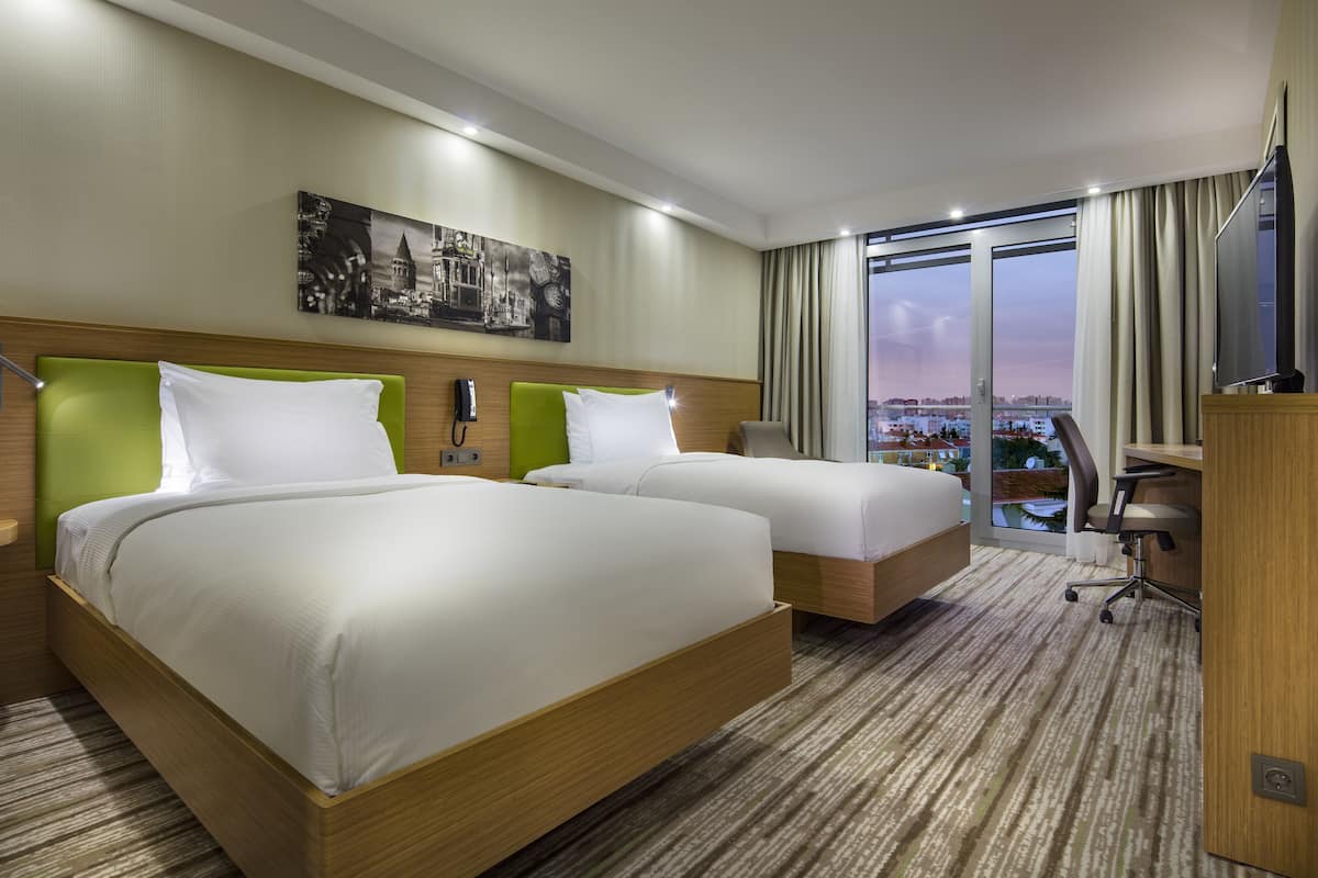 A hotel room with two beds, a desk, a chair, and a large window with a city view.