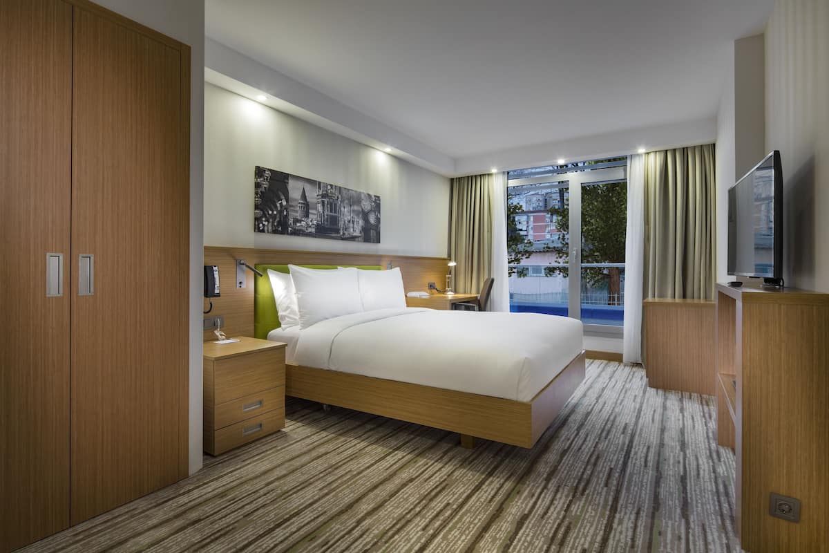 A modern hotel room with a large bed, wooden wardrobe, bedside table, lamp, and a TV.